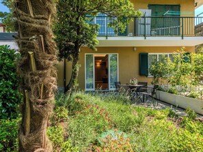 A two-story building with a balcony, a garden with various plants and flowers, and a dining area with outdoor furniture. - Nice apartment for 4 people with A/C and patio (Colico)