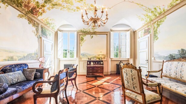 Una habitación con piso de parquet de madera, un candelabro, un sofá, una silla, un aparador y vistas a través de las ventanas.