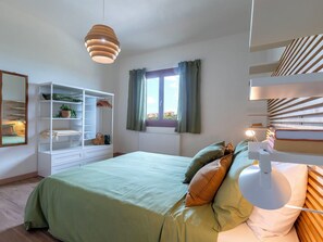 Ein Schlafzimmer mit Bett, Schrank, Pendelleuchte, einem Fenster mit Vorhängen und einem Nachttisch mit Lampe.