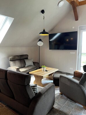 A modern loft-style living room with a high ceiling, a flat-screen TV mounted on the wall, a wooden coffee table, and a sofa with a checkered pillow. - Spacious penthouse apartment for all ages under one roof in an idyllic village setting with a sauna (Rubkow)