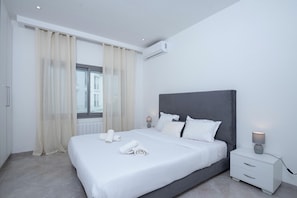 A modern hotel room with a large bed, bedside tables, an air conditioning unit, and a window with curtains.