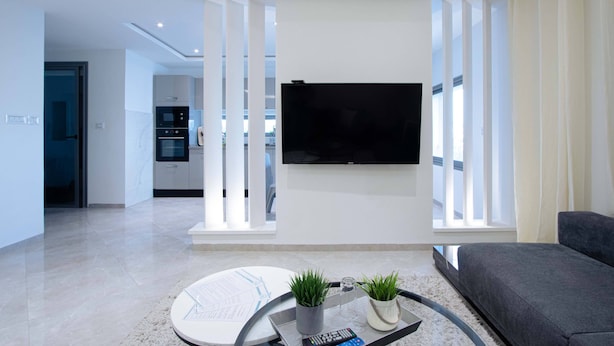 A modern living room with a flat-screen TV mounted on the wall, a grey sofa, a coffee table with a remote control, and a potted plant. - Sandra 5 (La Marsa)