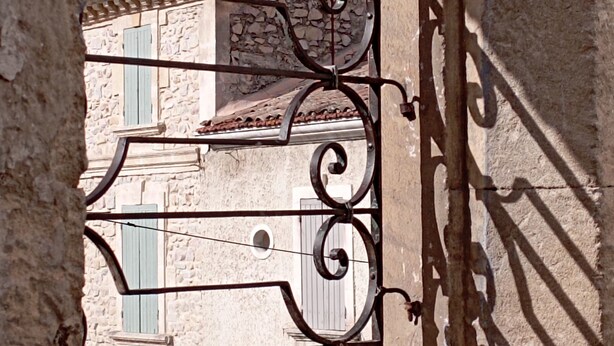 Un bâtiment en pierre, doté d’un toit en tuiles et de volets verts, vu à travers une arche ornée de motifs en métal.
