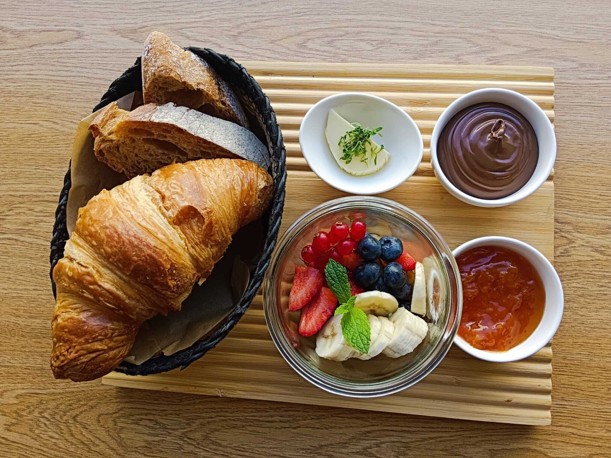 A wooden table with a basket of croissants, a bowl of mixed berries, and two small bowls of sauces.