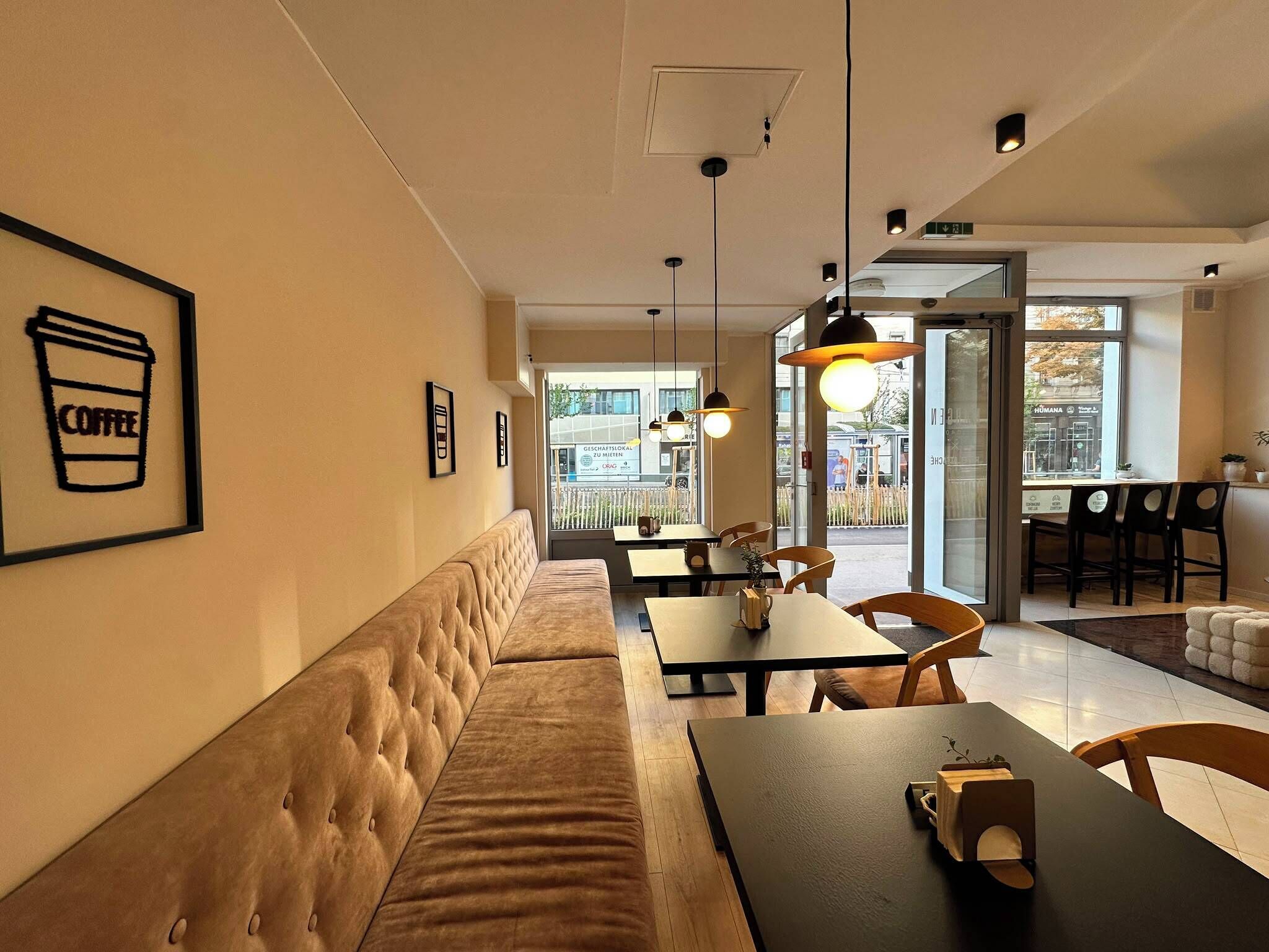 Interior moderno de cafetería con un largo banco acolchado, mesas y lámparas colgantes.