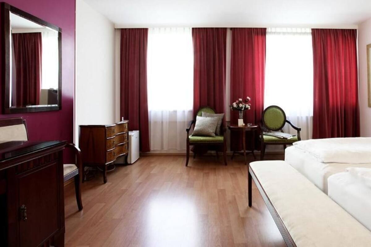 A hotel room with a wooden floor, a bed, a desk with a chair, a dresser, and a mirror.