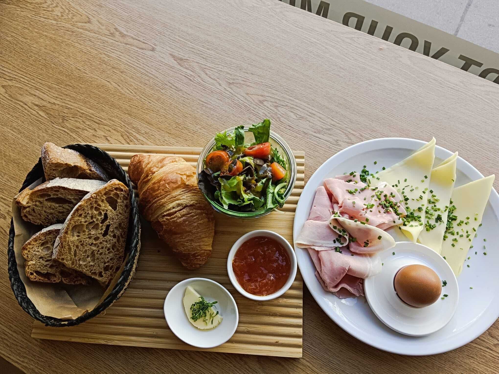 A wooden table with a variety of food items including bread, a salad, a plate of cheese and ham, and a small bowl of sauce.