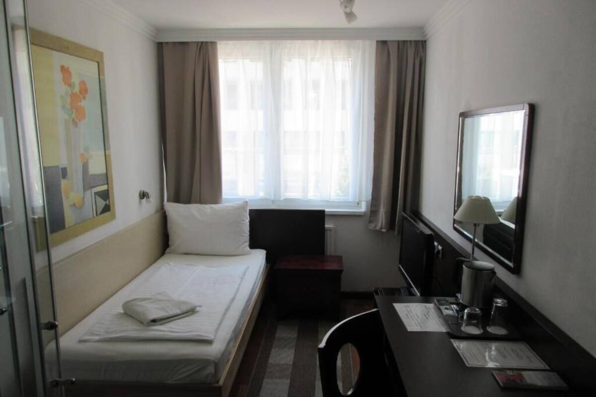 A hotel room with a bed, a desk, a chair, a TV, a window with curtains, and a painting on the wall.