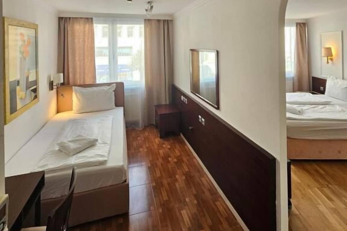 A hotel room with a wooden floor, two beds, a desk, a chair, a TV, and a window with curtains.