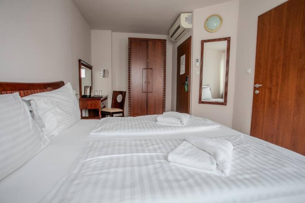 A hotel room with two beds, a wooden wardrobe, a desk with a chair, a mirror, and an air conditioning unit.