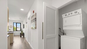 A modern kitchen with a built-in washing machine and dryer, a dining area with a view, and a hallway with artwork. - Exquisite 1BR 30 min to FIFA (Bayonne)