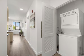 A modern kitchen with a built-in washing machine and dryer, a dining area with a view, and a hallway with artwork. - Exquisite 1BR 30 min to FIFA (Bayonne)