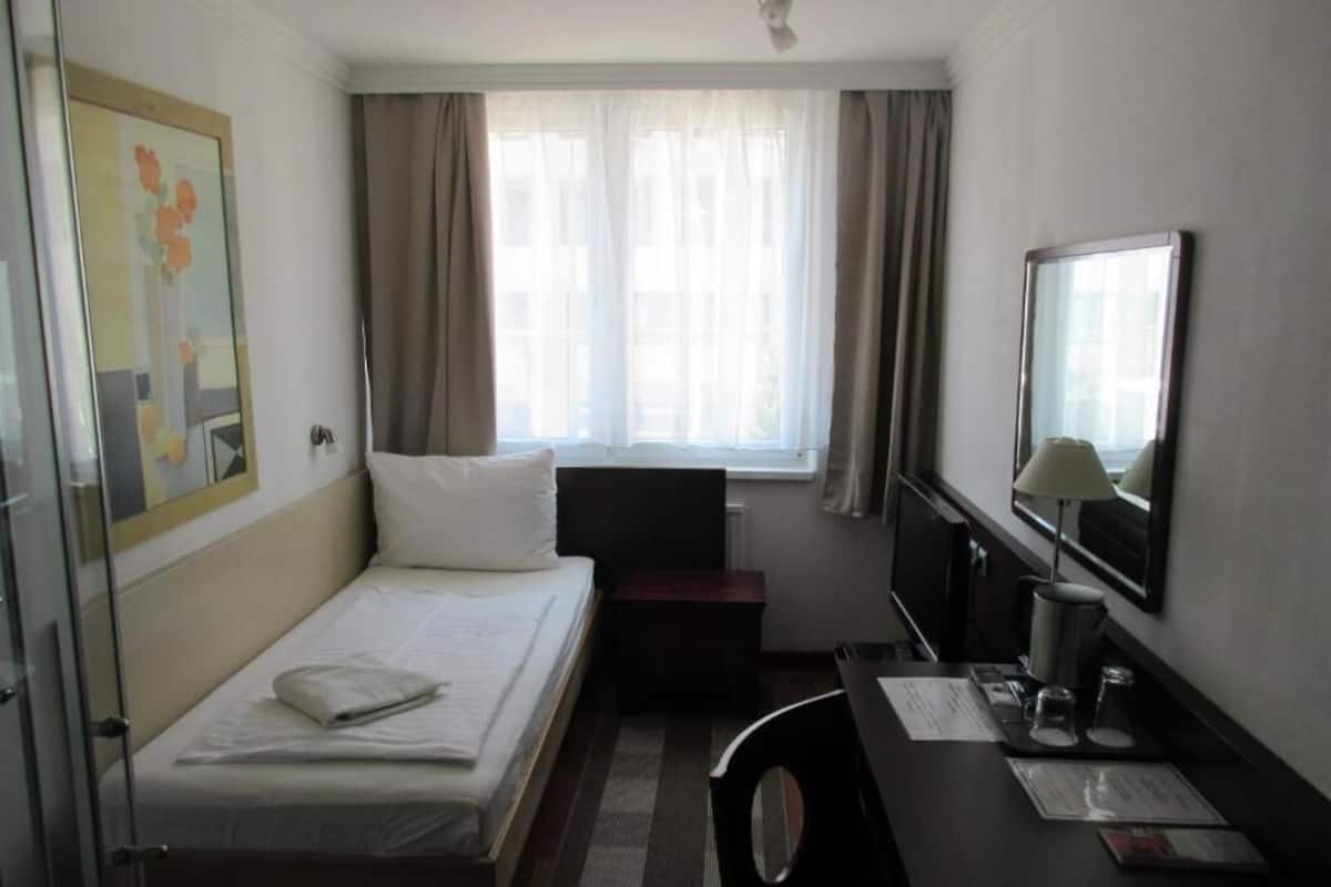 A hotel room with a bed, a desk, a chair, a TV, a window with curtains, and a painting on the wall.
