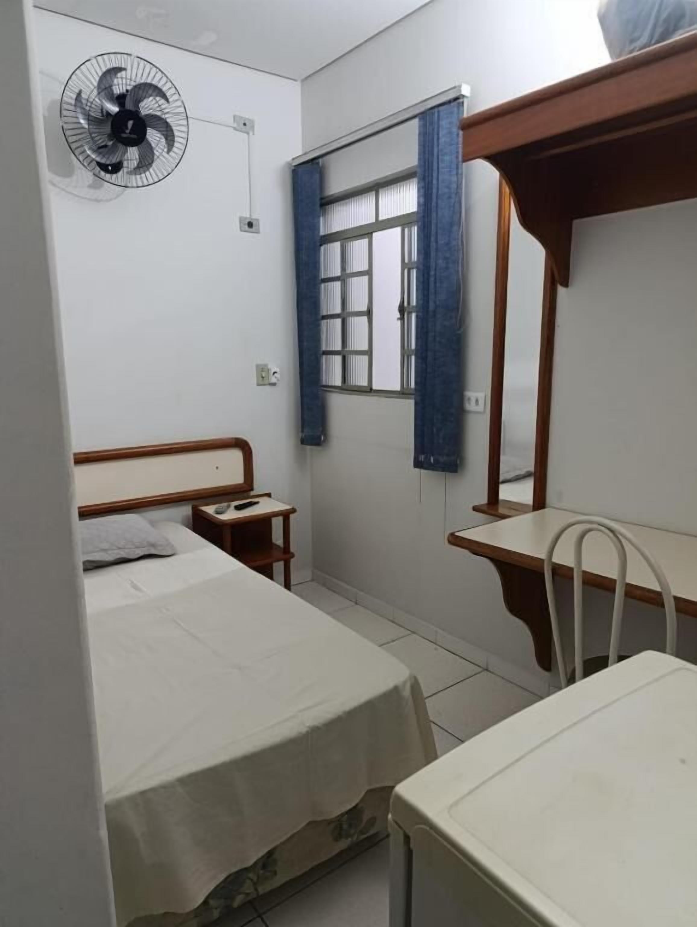Quarto com cama, mesa de cabeceira, ventoinha, janela com cortinas e escrivaninha com cadeira.