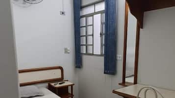Quarto com cama, mesa de cabeceira, ventoinha, janela com cortinas e escrivaninha com cadeira.