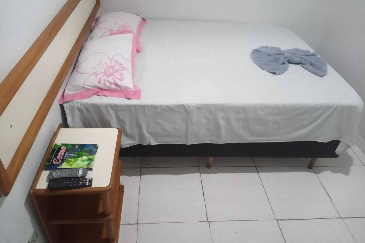 Uma cama de solteiro com colchão branco e almofadas cor-de-rosa, cabeceira de madeira, mesa de cabeceira pequena com um controle remoto e um cobertor cinza sobre a cama.