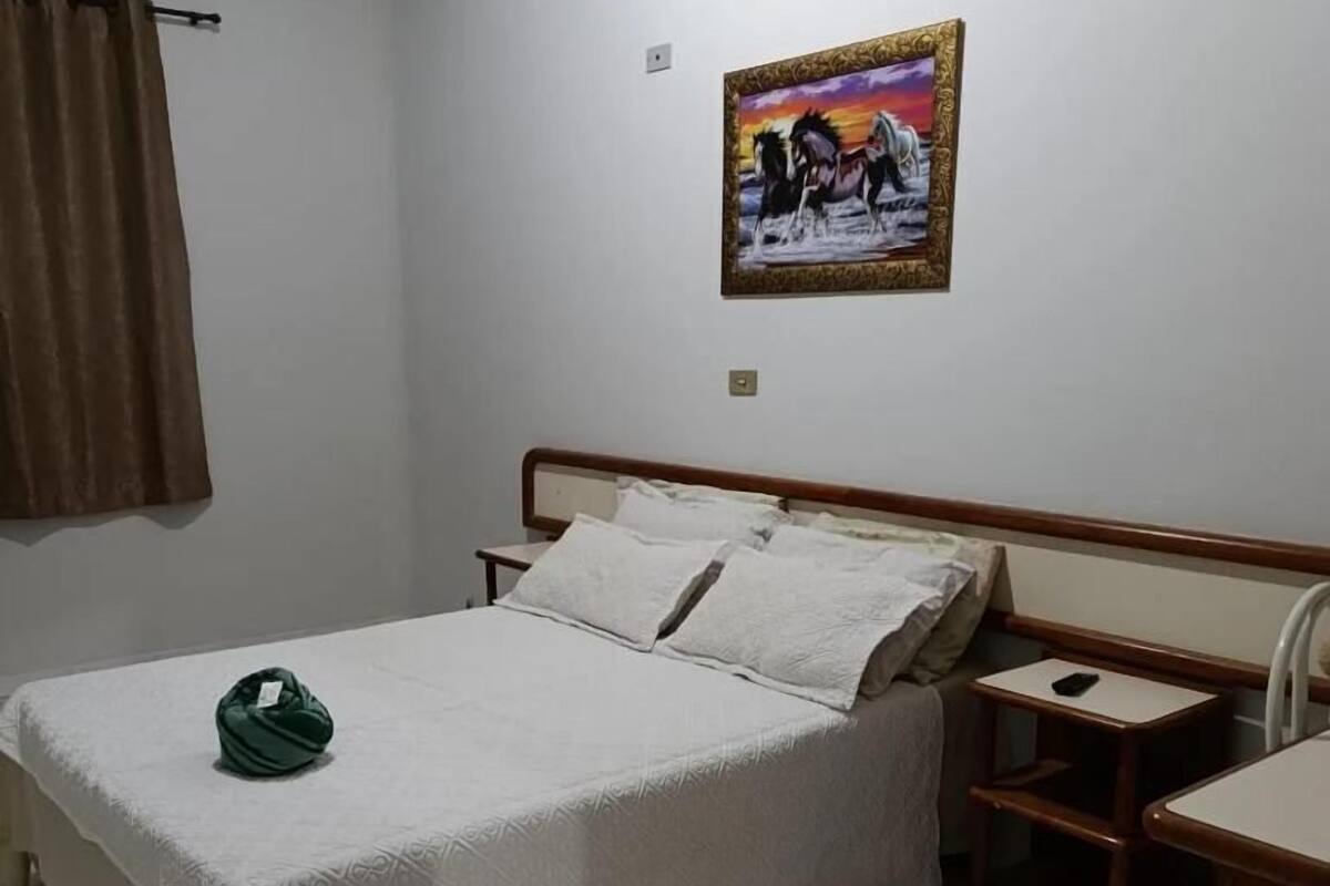 Quarto com cama, mesa de cabeceira, cadeira, mesa e um quadro na parede.