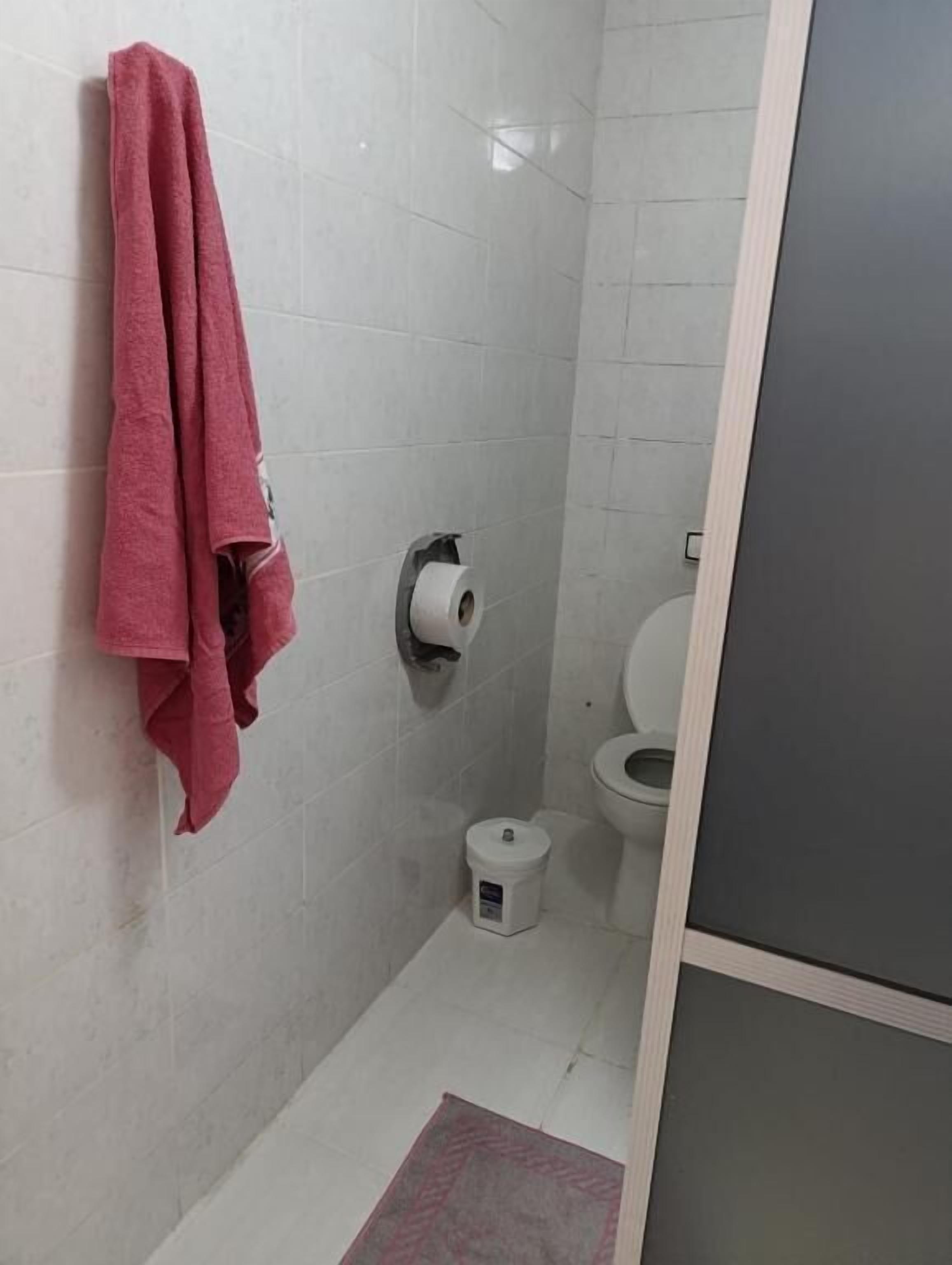 Een kleine badkamer met een toilet, een handdoekrek met een handdoek, een toiletrolhouder en een prullenbak.