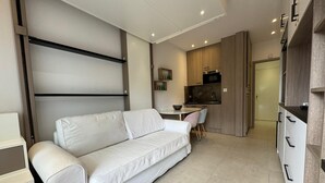 Una sala de estar moderna con un sofá blanco, una pequeña mesa de comedor y una zona de cocina con armarios de madera y un microondas.