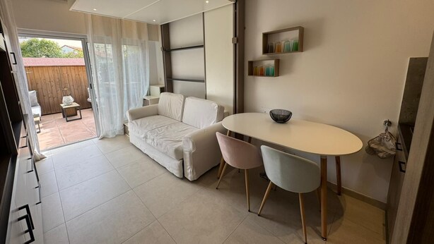 Una sala de estar moderna con un sofá, una mesa de comedor con sillas, una puerta corrediza de vidrio que da a un patio exterior y un estante pequeño con libros.