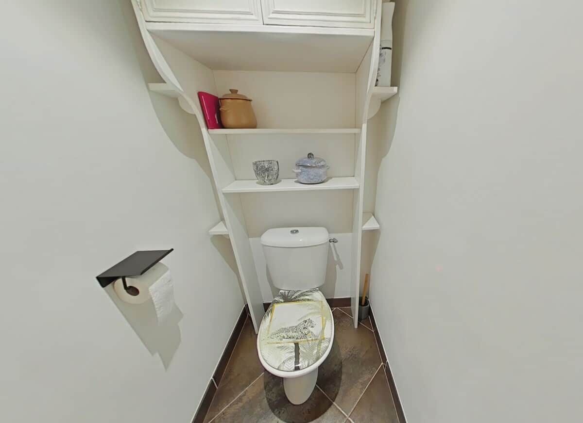 A small bathroom with a toilet, a shelf with a pot and a container, and a roll of toilet paper.