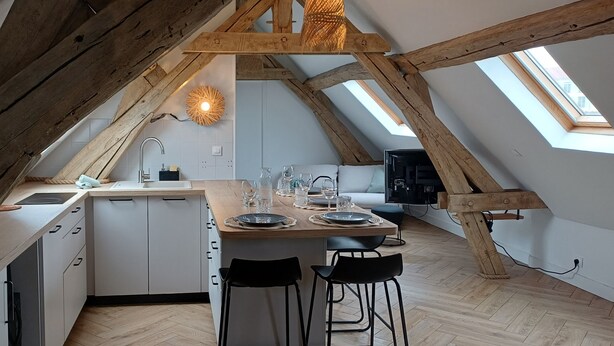 Une cuisine de style industriel, avec un plafond orné de poutres en bois, un puits de lumière, des armoires blanches et un îlot central doté de tabourets de bar.