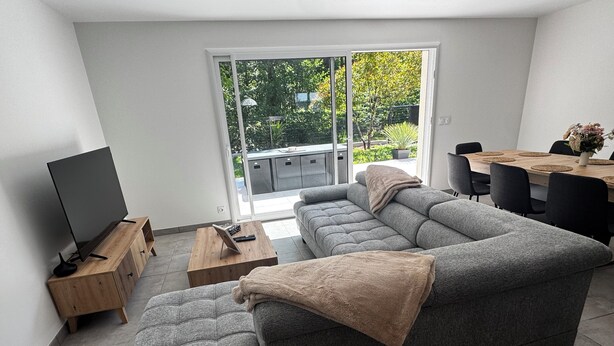 A modern living room with a grey sectional sofa, a wooden coffee table, a flat-screen TV, and a dining area with a wooden table and chairs.