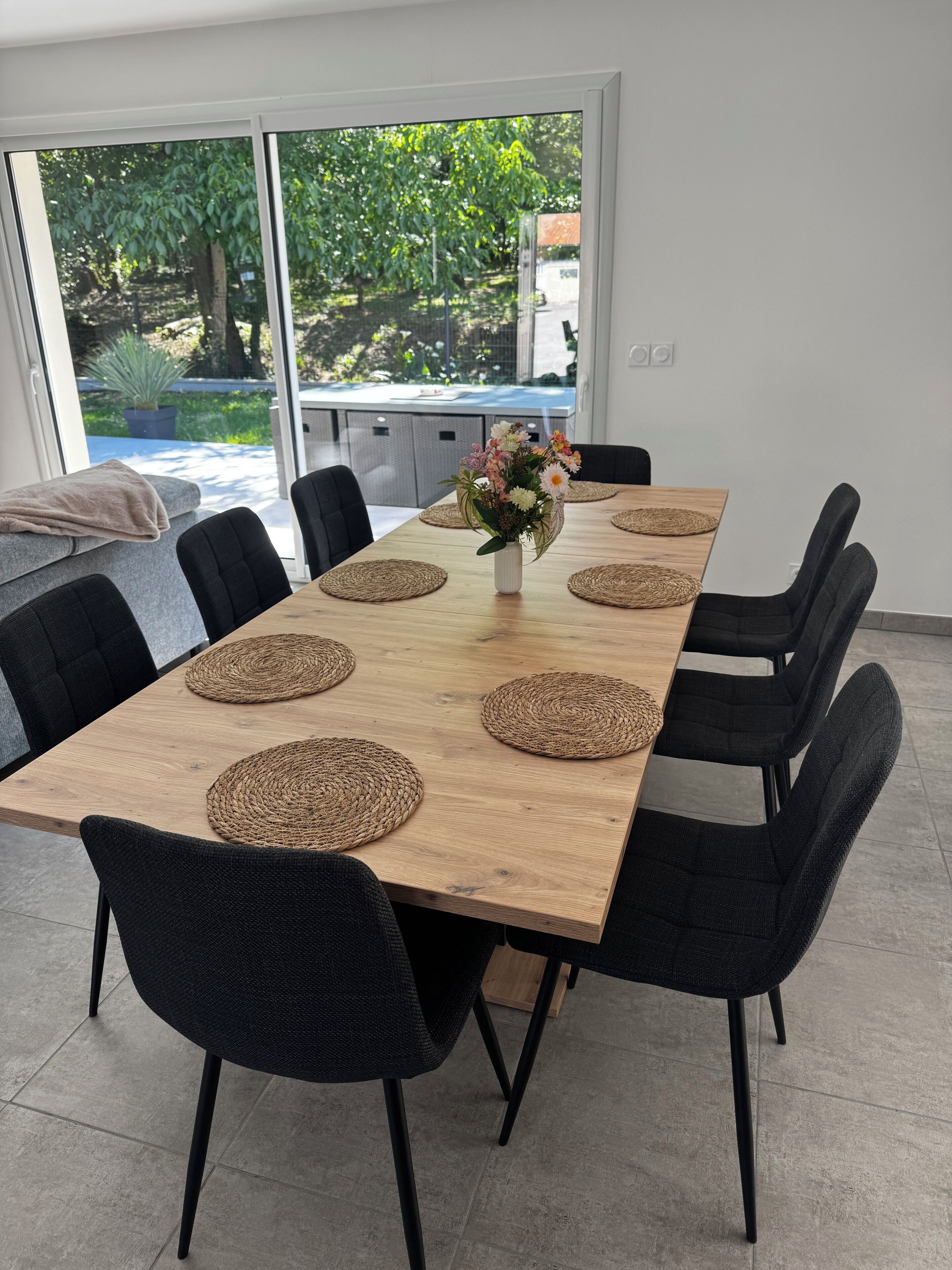 Ein Essbereich mit einem langen Holztisch, schwarzen Stühlen, gewobenen Tischsets und einer Vase mit Blumen.
