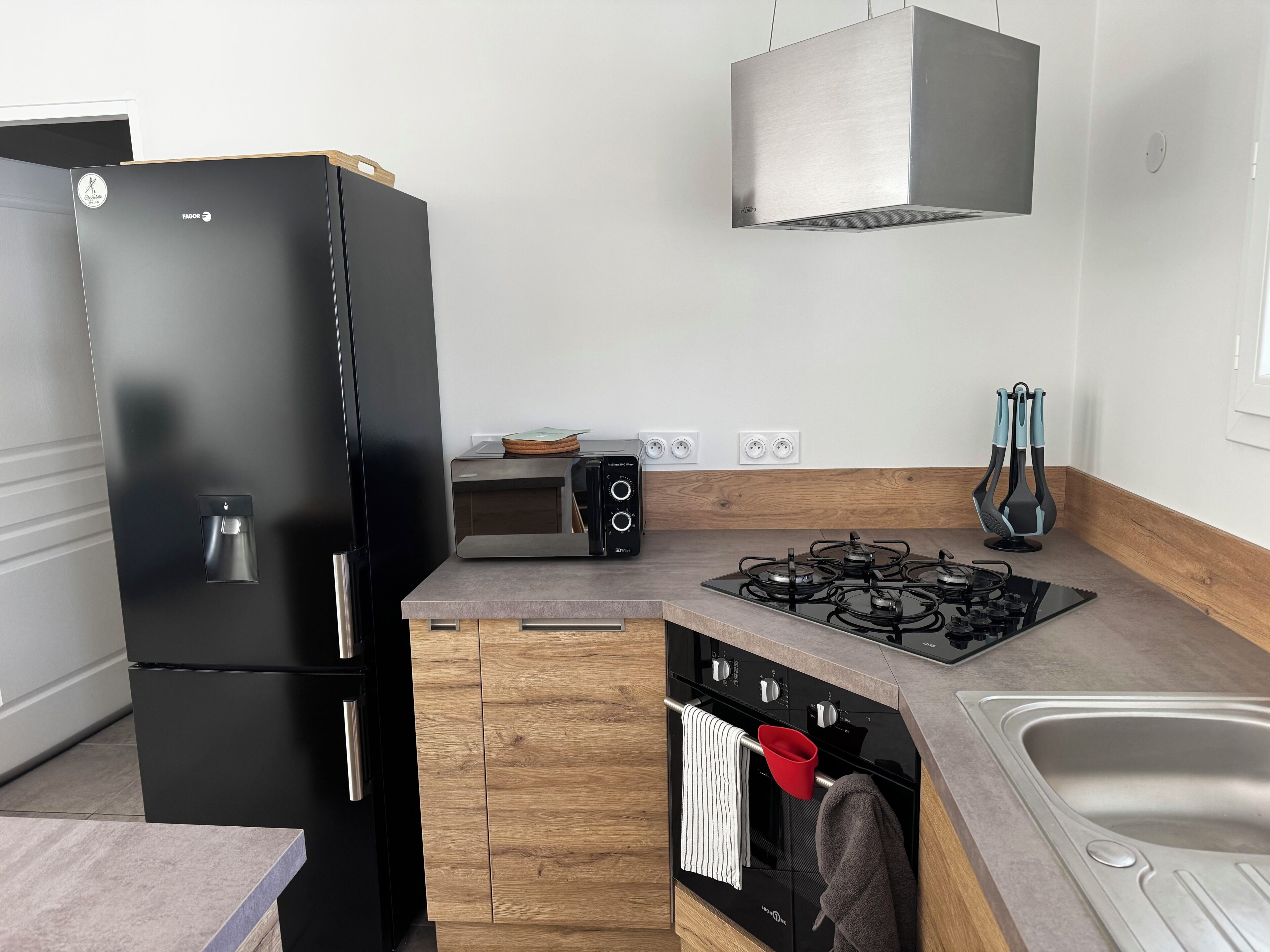 Una cuina moderna amb un refrigerador negre, un forn integrat, una placa de gas i un rentaplatets d'acer inoxidable.