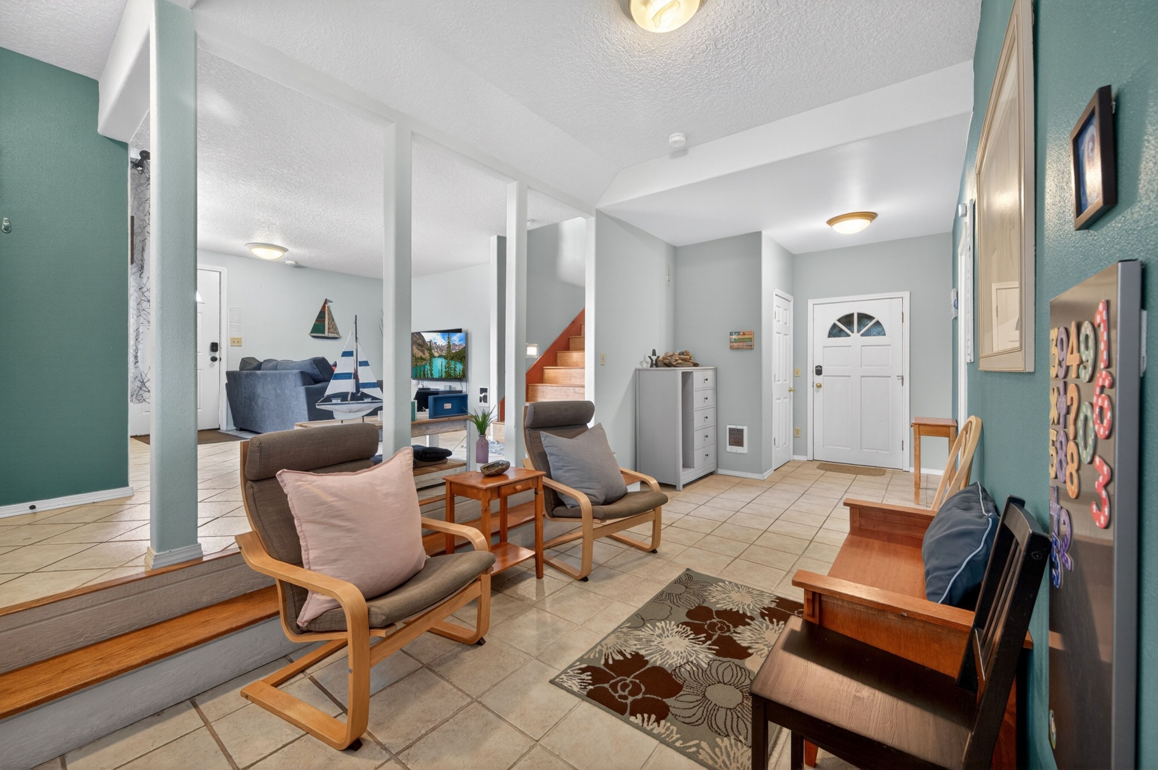 A spacious living area with a sofa set, a coffee table, and a rug. There is a staircase leading to another level, and a door with a number display.