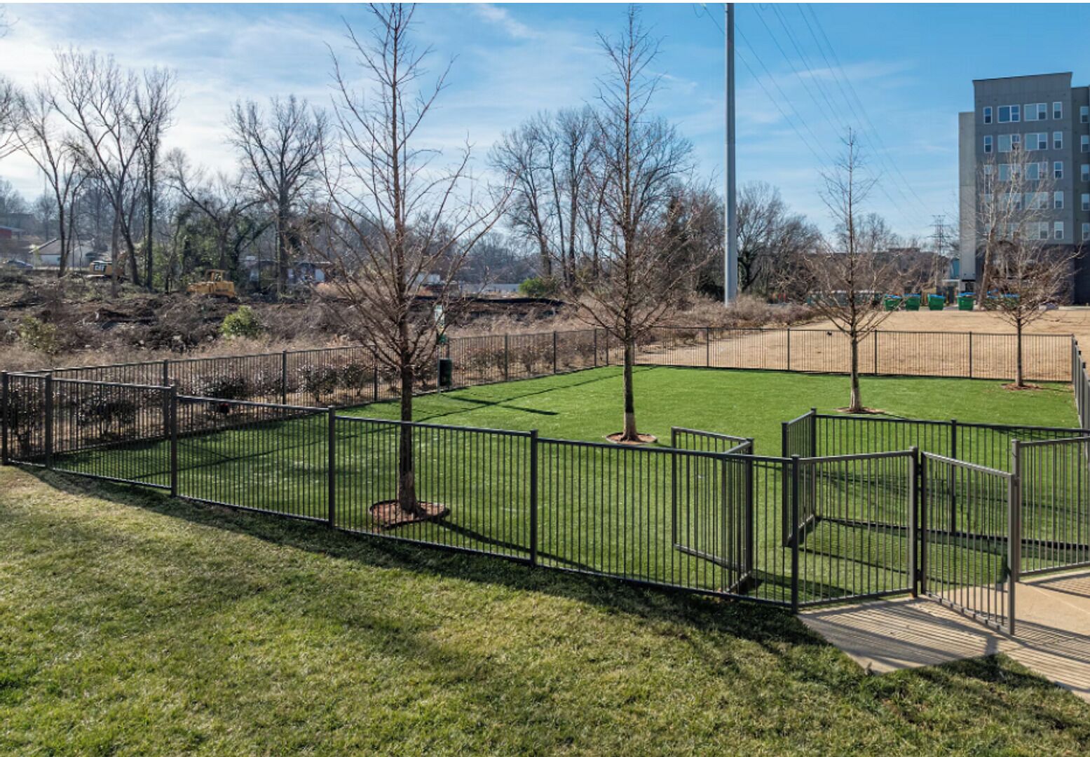 A fenced park area with grass and trees, a building in the background, and a clear sky.