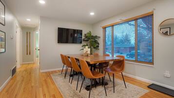 A dining area with a wooden table and chairs, a potted plant, and a large window with blinds.