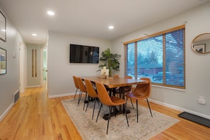 A dining area with a wooden table and chairs, a potted plant, and a large window with blinds. - Central Gem ~ Backyard Oasis with Hot Tub and Firepit (Arvada)