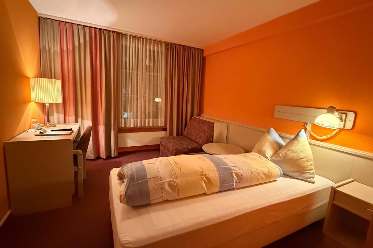 Una habitación de hotel con cama, escritorio, silla y ventana con cortinas.