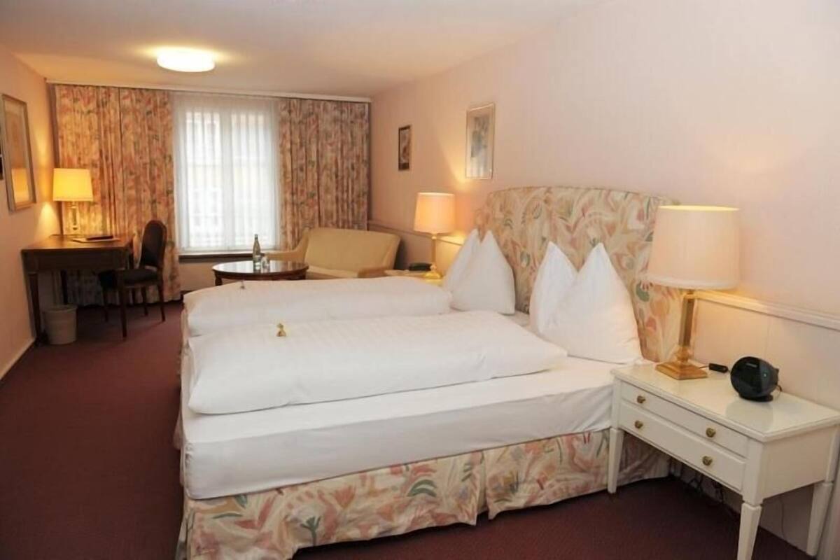 Habitación de hotel con cama doble, escritorio, silla, dos mesitas de noche con lámparas y ventana con cortinas.