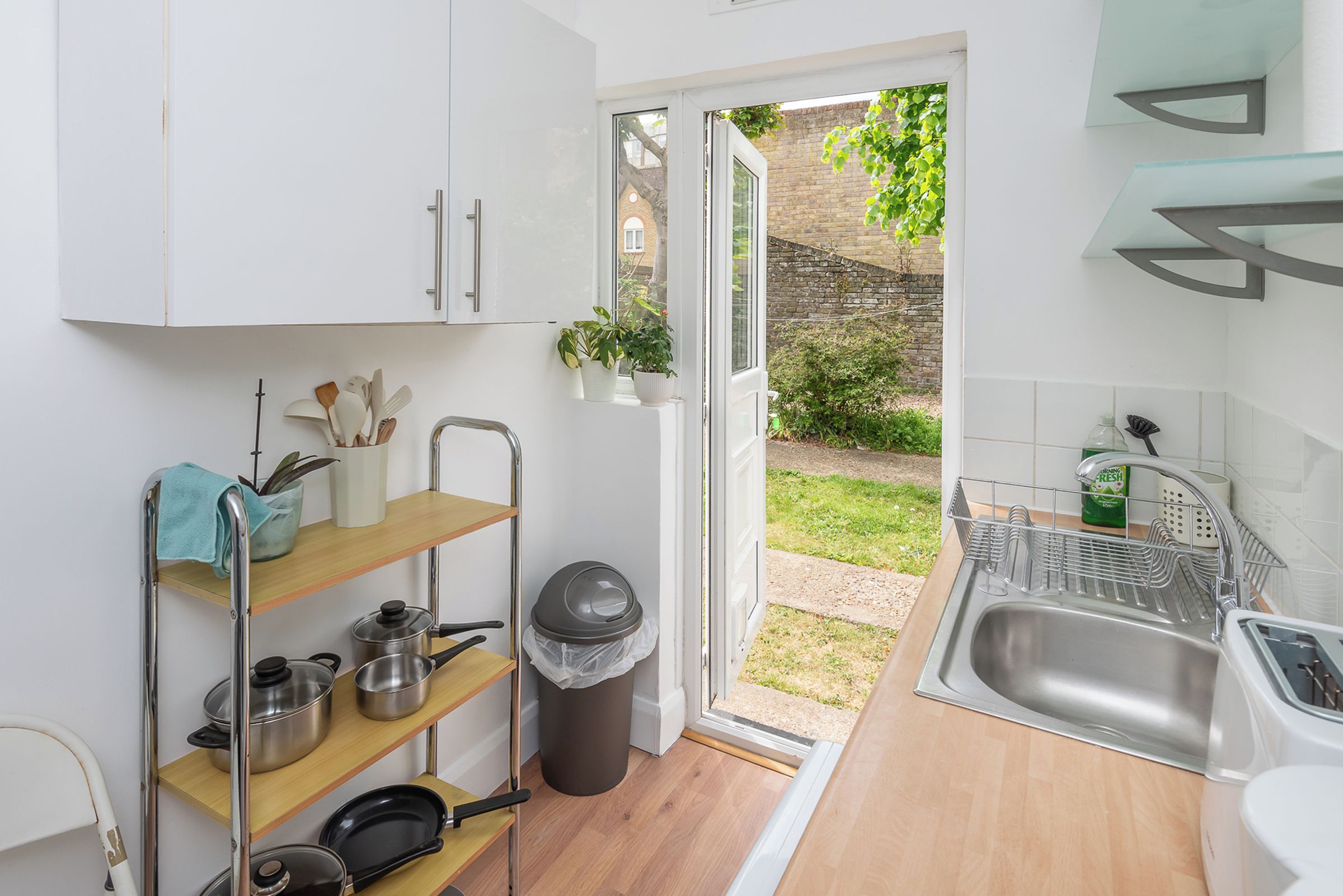 A compact kitchen with white cabinets, a wooden shelf, and a view of a garden through an open door.