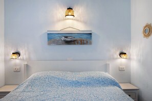 A bedroom with a bed, two wall-mounted lamps, a painting of a boat on water, and a wall sconce.