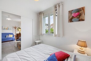 A bedroom with a bed, a nightstand, a lamp, a window with curtains, a painting, and a view of the outside. - Holiday Home 'Gîte « le Bel Air »' with Private Terrace, Private Garden and Wi-Fi (Saint-Gilles-Croix-de-Vie)