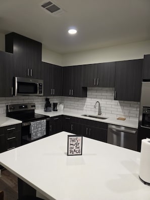 A modern kitchen with dark cabinets, a stainless steel microwave, oven, and dishwasher, a white countertop, and a sign that says 'THIS MUST BE THE PLACE'. - Cozy 1-bedroom apartment in charming Addison (Addison)