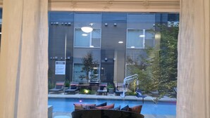 Janela com vista para área da piscina, mobiliário de exterior e um edifício com várias janelas e um letreiro.