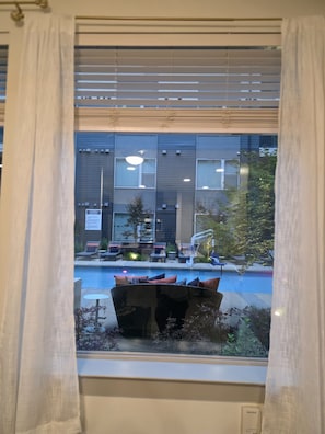 A window with a view of a pool area, outdoor seating, and a building with multiple windows and a sign. - Cozy 1-bedroom apartment in charming Addison (Addison)
