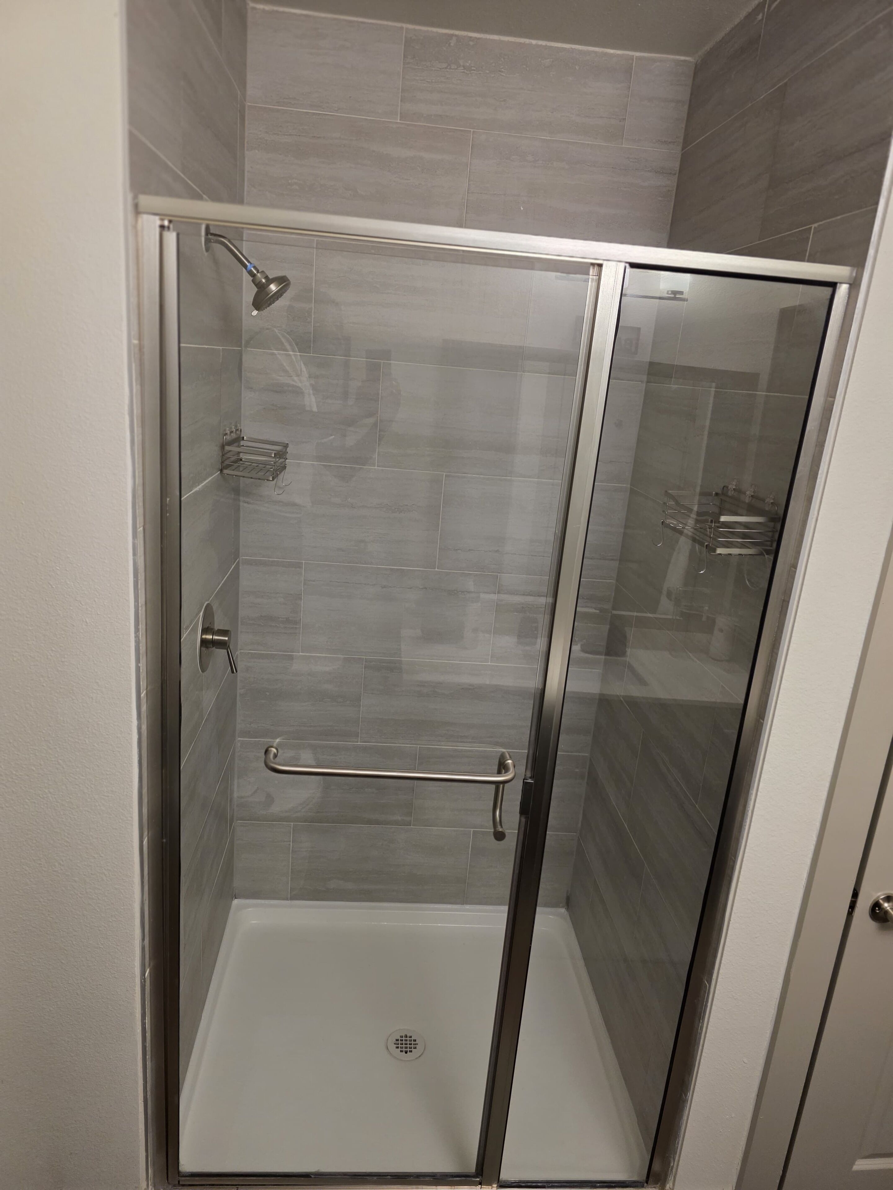 A glass-enclosed shower stall with a metal grab bar, a white tiled floor, and a wall-mounted showerhead.