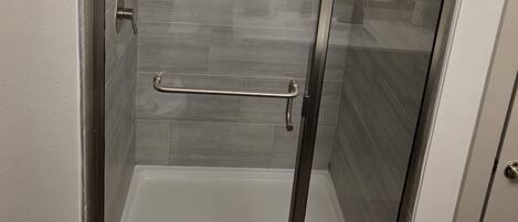A glass-enclosed shower stall with a metal grab bar, a white tiled floor, and a wall-mounted showerhead.