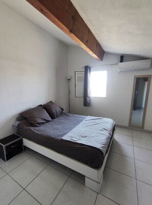 A bedroom with a bed, a nightstand, a window with curtains, an air conditioning unit, and a door. - Villa Torreilles Plage 4 People (Torreilles)