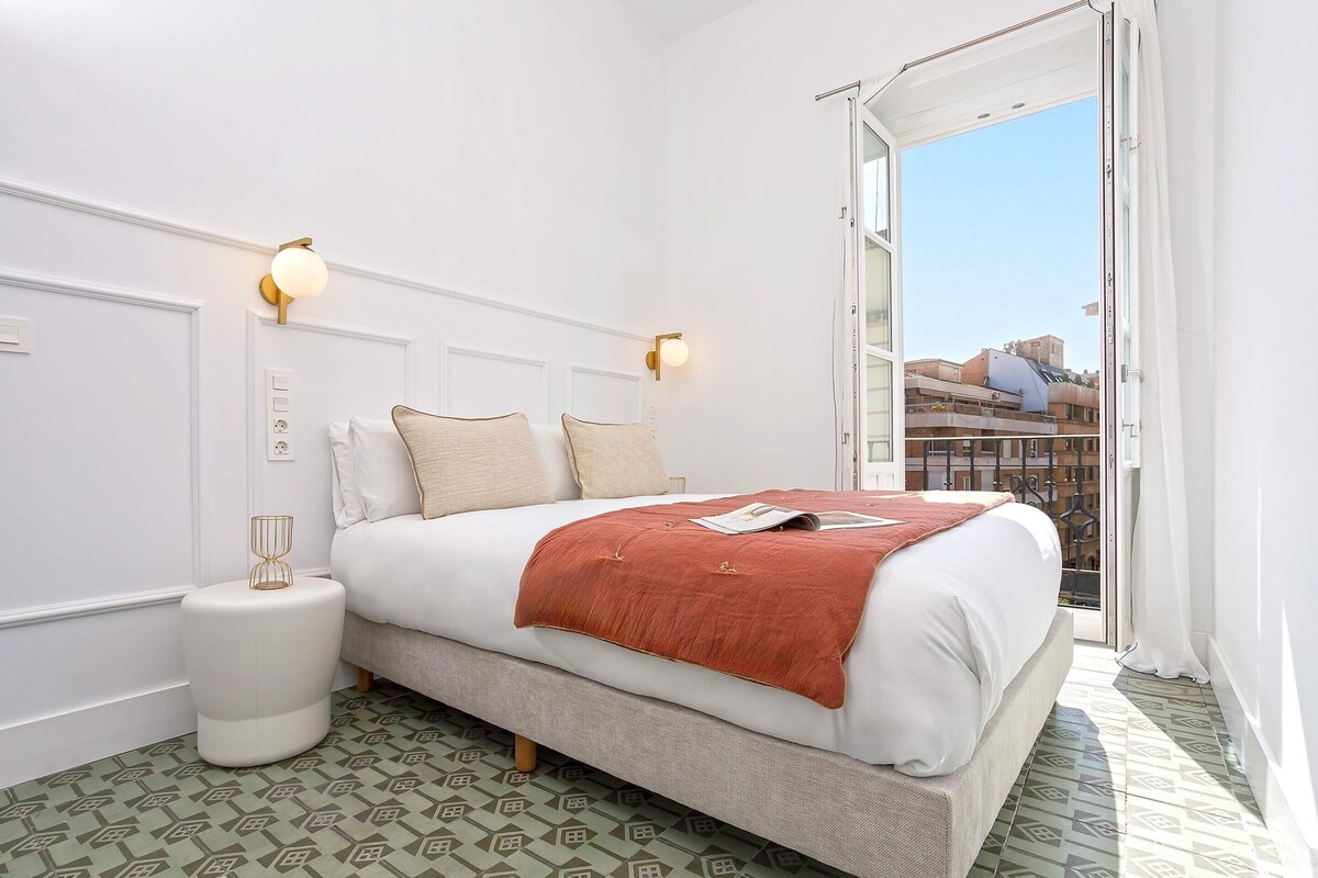 Habitación de hotel con cama, lámparas de noche, una mesita y vistas a edificios a través de la ventana.