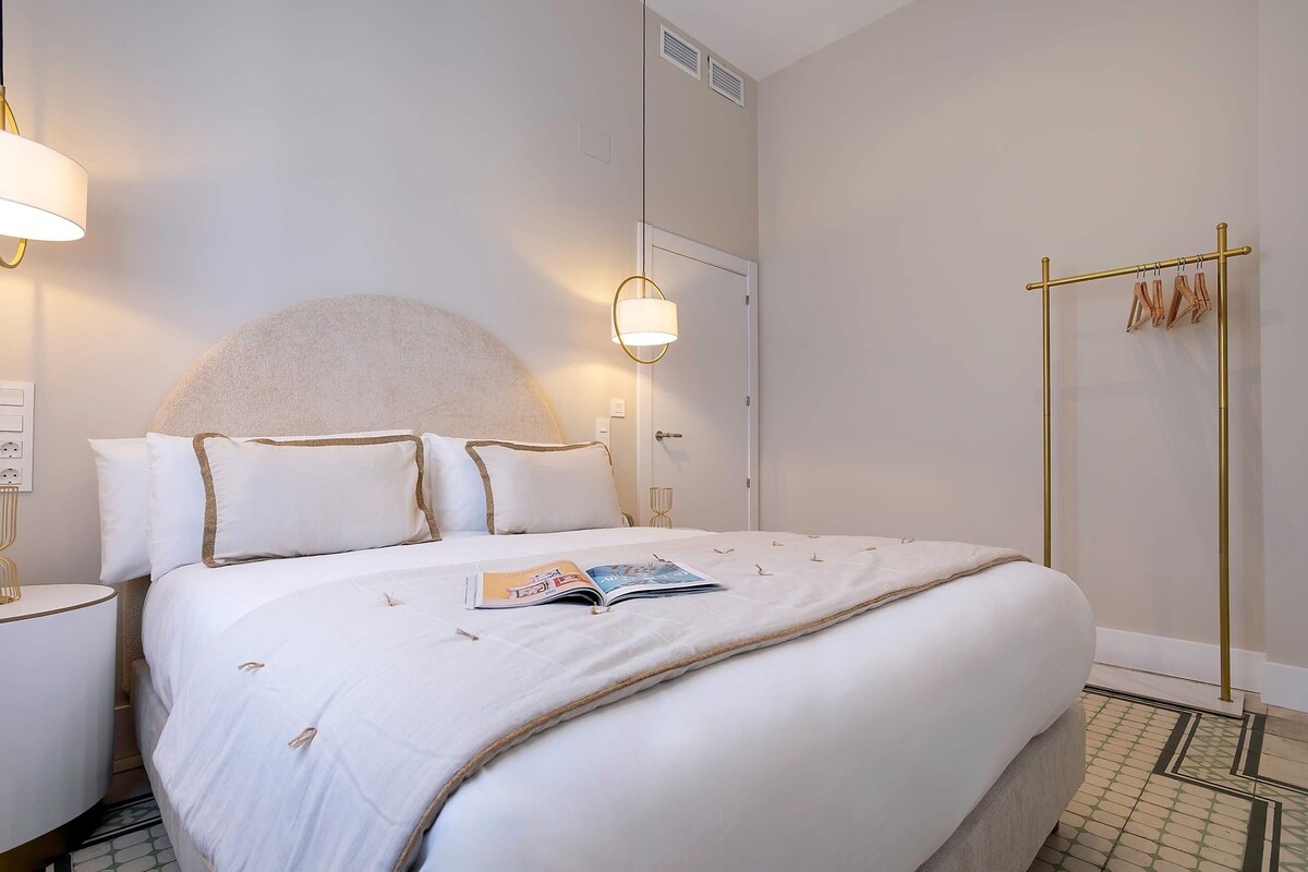 Una cama bien hecha con una revista, una lámpara de noche y un perchero con ropa.