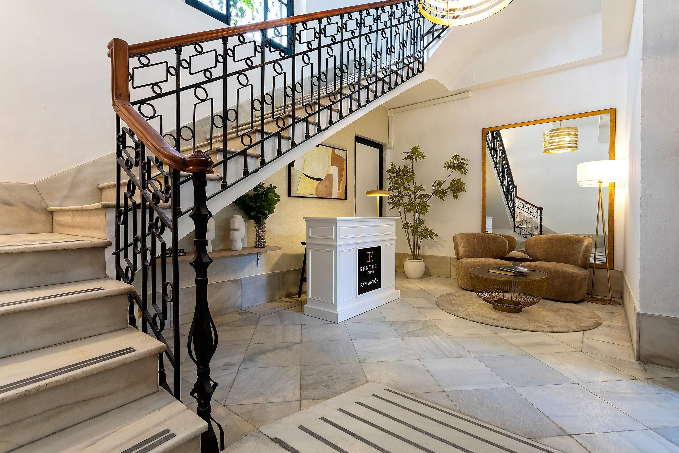 Una escalera con barandilla de hierro forjado que conduce a una plataforma, una chimenea blanca, un sofá y una mesa de centro en un salón.