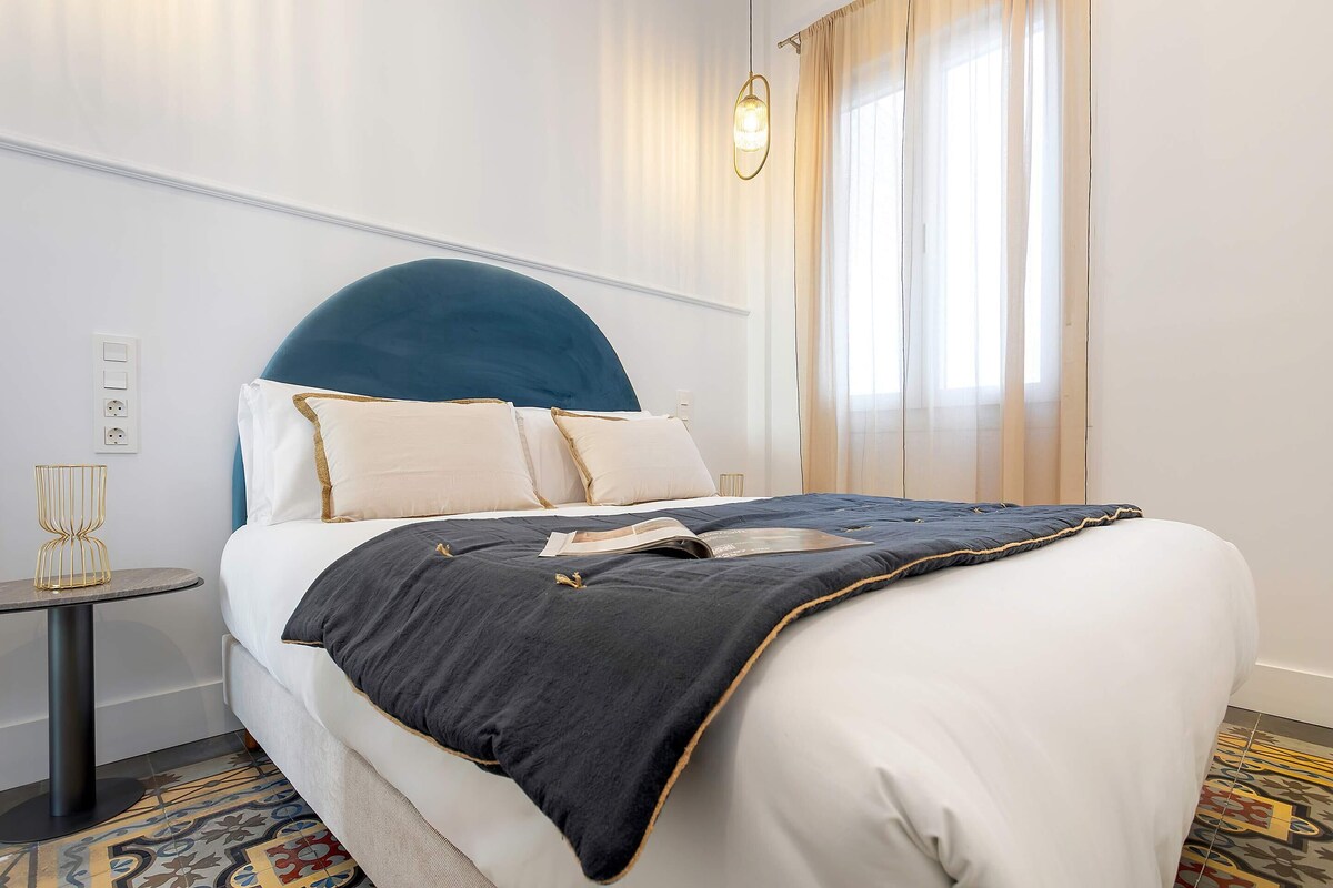 Una cama bien hecha con un cabecero oscuro, ropa de cama blanca y una alfombra estampada.