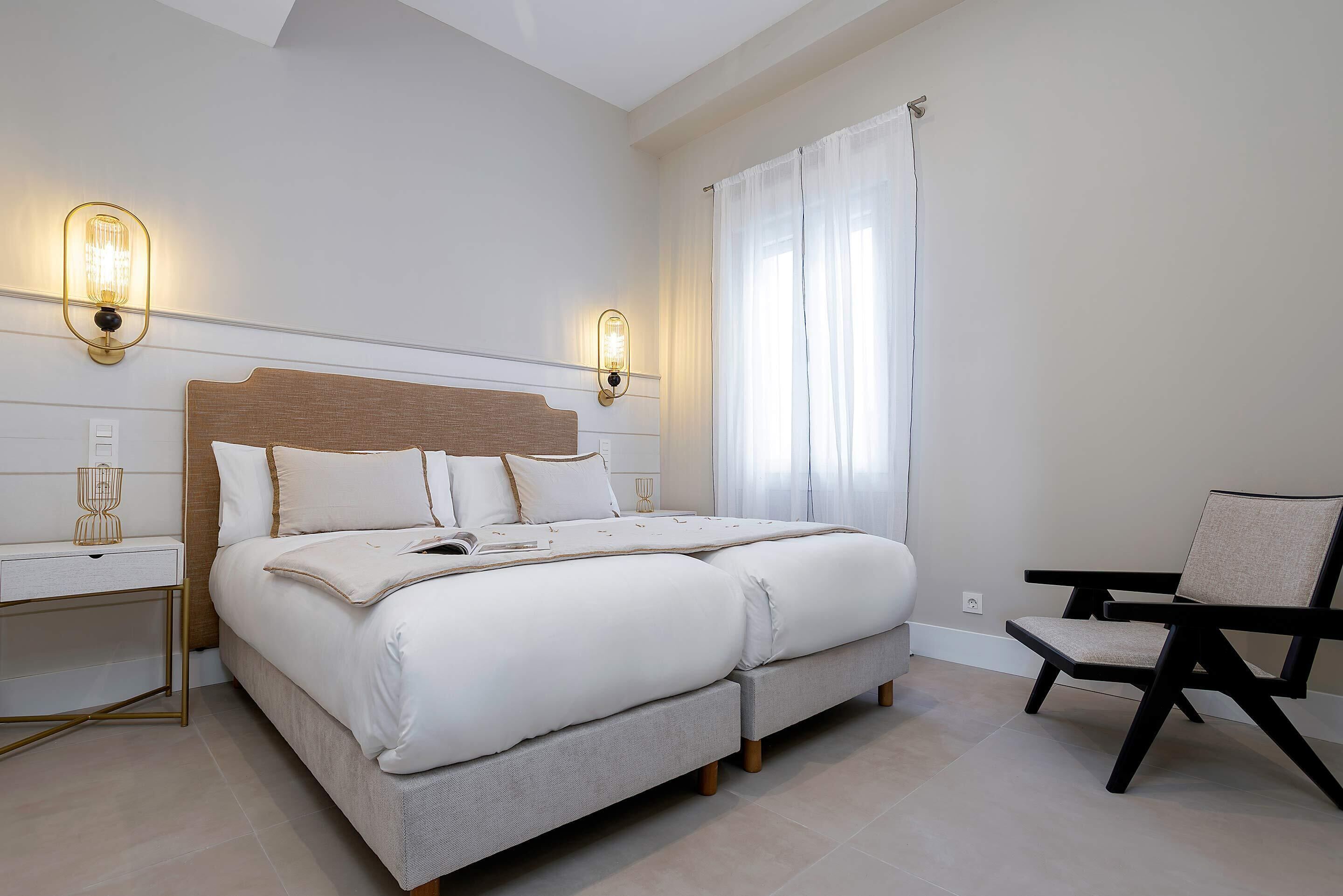 Un dormitorio con una cama grande, mesitas de noche, una silla y dos lámparas de pared.