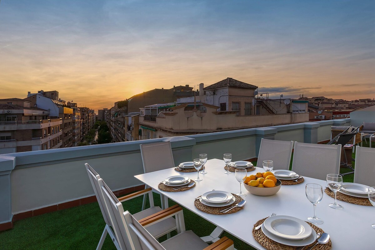 Un área de comedor en la azotea con una mesa dispuesta para cuatro personas, con vista a un paisaje urbano al atardecer.